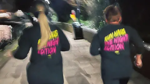 Two women are running at the Lakeside in Doncaster. They wear shirts reading "Run Haus: The Night Edition" on the back.