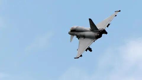 PA A grey fighter aircraft is seen in the distance flying against a blue sky 