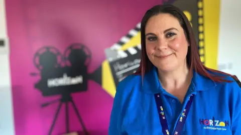 Lucy smiles for the camera. She is wearing a bright blue t-shirt with the Horizon logo and stands before a mural with a filming theme.