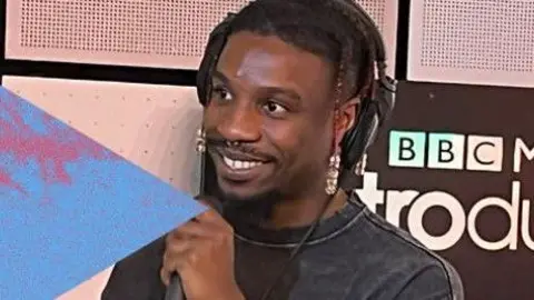 Native James wearing a black T-shirt and headphones is smiling and holding a microphone.