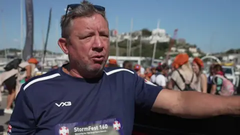 Richard Burden is wearing a short sleeved blue top with the words Stormfest 160 on it.
Members of the gig crew are in the background with their orange mob caps.