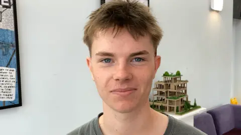 Luke Deal/BBC Toby Groom, who has short brown hair and is wearing a green t-shirt, smiles at the camera