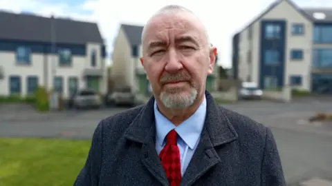 A grey haired man with a grey coat and red tie and blue shirt on 