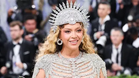 US performer Beyonce poses on the red carpet for the 2026 Met Gala wearing a shimmering skeletal gown and a spherical headpiece as photographers in suits stand behind her.