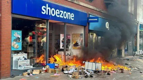 Shoezone shop with items strewn and ablaze on the pavement in front of the large store window.