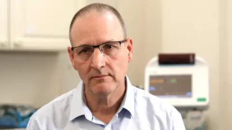 Martin Rolles looks at the camera. He wears a white shirt and glasses, and short dark hair. Medical equipment with a screen can be seen behind him. 