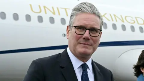 PA Media Sir Keir Starmer arriving at a NATO summit, walking on airport tarma near a United Kingdom aircraft. A uniformed officer stands beside the stairs leading up to the plane.