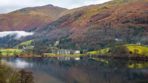 AFP via Getty Images The village of Grasmere as seen from across the lake. Mist hangs over the trees and fells rise in the background.