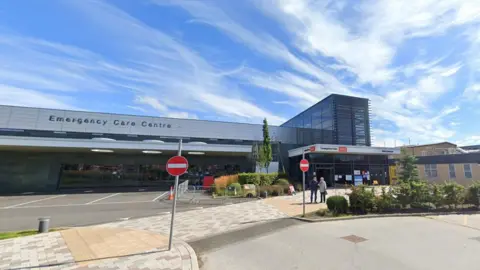 Google A&E at Queen Elizabeth Hospital, Gateshead. It is a sunny day. The photo shows a grey building with the sign "Emergency Care Centre".