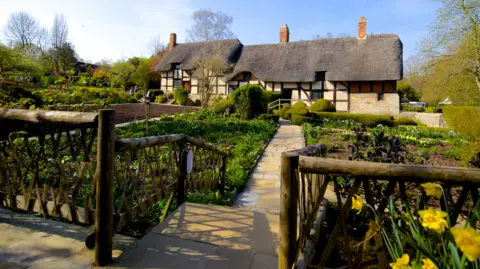 The Shakespeare Birthplace Trust Anne Hathaway's Cottage is in the background, with gardens in the foreground and to the left of the cottage on the photo. A walkway goes from the bottom of the image in the middle to the cottage.