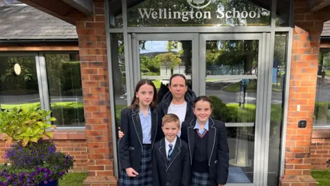 BBC Rebecca with her three children, all wearing school uniforms, standing outside the entrance to Wellington School