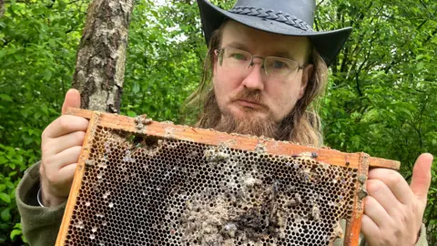 Lee holds up a frame from an old beehive. It is blackened and appears old, with several bees still stuck to the mesh of the frame.