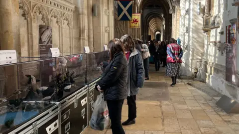 Tom Jackson/BBC A long run of glass cases containing the display line a cathedral wall, with two women looking at it. The cathedral is busy, with people gathered at the back of the photo.