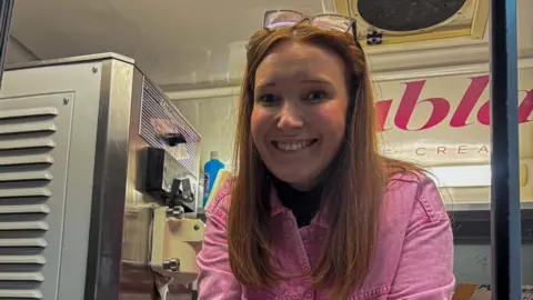 Lauren Evans leaning out of ice cream shop window wearing a pink shirt. She has red long hair and glasses pushed up into her hair