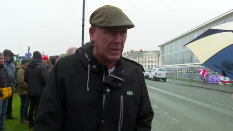 Gareth Wyn Jones lasting  the otherside of the roadworthy  from Venue Cymru, wherever  the Welsh Labour league  is taking place. He is wearing a level  headdress  and a rainfall  overgarment  and determination   are different   protesters down  him and the entity  is grey