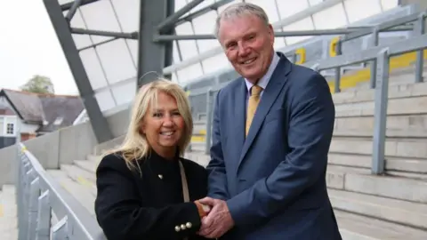 Leeds Rhinos Gill Spencer stands close to Andrew Thirkill in the stand of a stadium. The pair are locked in a handshake and smiling into the camera. Gill is wearing a smart black coat with brass buttons while Andrew is in a blue suit and yellow tie.