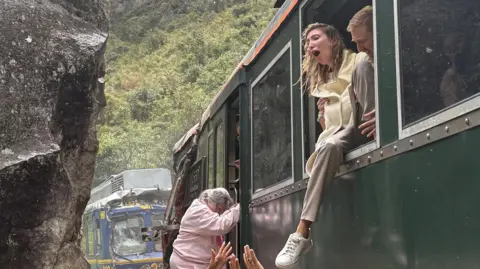 Reuters A woman screams as she exits a crashed train through the window. Others beneath her try to help, reaching up towards her. There is a clear sense of panic on their faces. Behind them, a crashed train and a large cliff. 