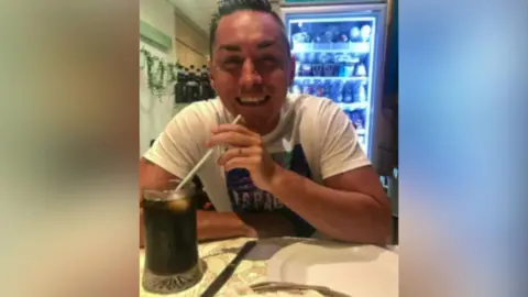 Jamie Attwood with short dark hair smiling at the camera and wearing a white T-shirt with a logo on the front. He is holding a straw which is in a glass full of a dark liquid.  There is a fridge behind him.