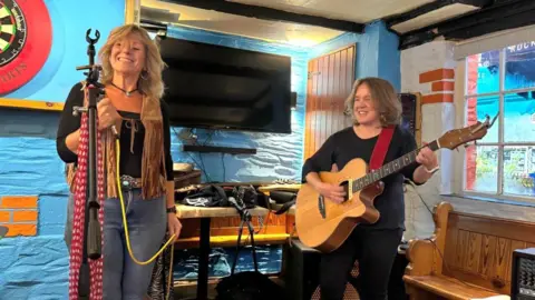 Penny Bulkeley Two women standing at the front of a pub performing. The woman on the left is smiling and is holding a microphone. The woman on the right is playing an acoustic guitar. The building's walls are blue. A TV is displayed on the wall behind them.