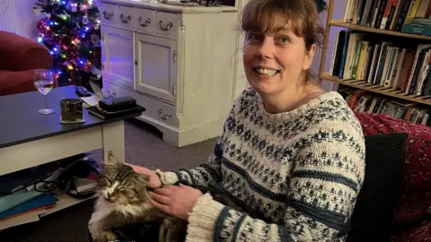 Claire Berry wearing a knitted jumper with brown hair in a pony tail stroking her cat, Rhubarb, who is sitting on her knee with a Christmas tree in the background