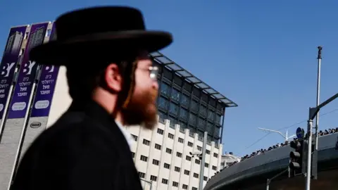 An ultra-Orthodox Jewish man pictured in the foreground, with a large concrete and glass building in the background (30/10/25)