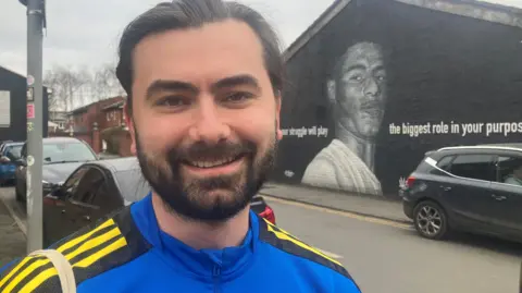 Larry Harizanov: a dark-haired man with a beard, wearing a blue jacket, standing in front of a Marcus Rashford mural.