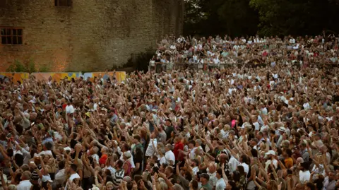 A large crowd of people outdoors, all facing the same way with a stone castle wall in the background