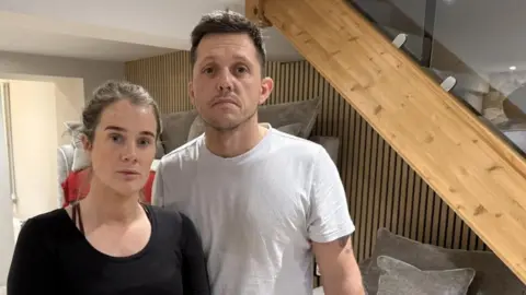 An unhappy looking man and woman standing in their living room which has been damaged by a flood.