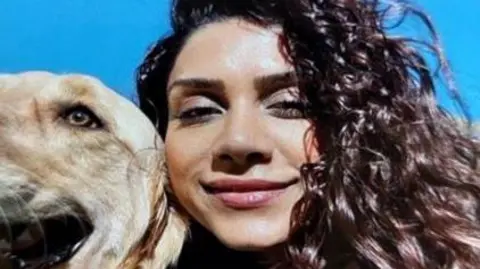 A close up selfie of Paria with a blonde golden retriever dog on a sunny day. She has brown curly hair and is smiling.