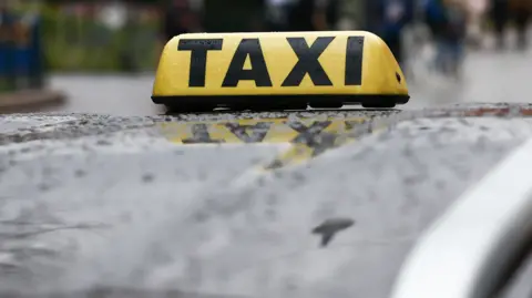 A close up of a sign saying taxi on top of a car. The letters are in black and the sign is yellow. The car is black and rain is on the roof. The background is out of focus. 