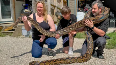 A man, a woman and a teenage boy all dressed in black and are kneeling down, each holding part of a large python. It has black, yellow and brown scaly skin. The family are knelt in front of a house and garage holding the snake.