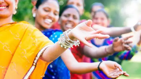 Bollywood dancers wearing colourful saris line up in a performance, with their arms outstretched making dance-themed hand gestures.