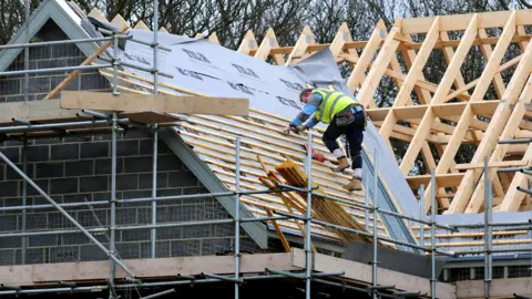 PA Media A builder wearing a hi-vis jacket works on a half-finished roof of a house surrounded by scaffolding
