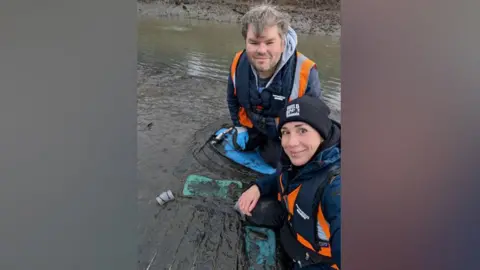 Dr Martina Bristow and engagement officer Blair Watson take a selfie during seagrass planting at the River Don near Jarrow. They are wearing high visibility blue and orange vests as they sit in the mud next to a frame which holds the young seagrass plants. Dr Bristow has brown eyes and a pierced nose and lip. She is wearing a black beanie. Blair Watson has short grey hair and blue eyes. 