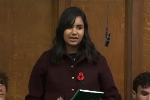 Sienna Ahir is seen standing in the House of Commons against a panelled background. She has a microphone hanging in front of her and is wearing a dark cardigan with gold buttons and a knitted poppy. 
