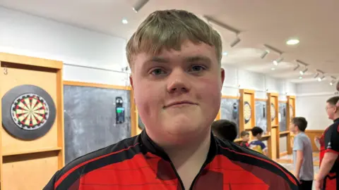 Brady is smiling - he has short blonde hair and is wearing a red and black sports top. Behind him is a hall lined with darts boards with young people throwing darts. 