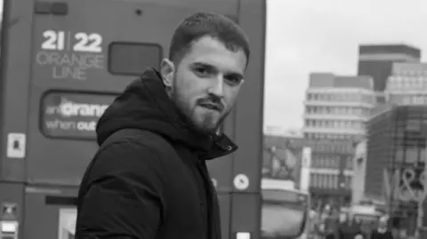 A black and white photo of a young man which short hair and a beard. He's stood in the middle of a road with a bus and busy street in the background