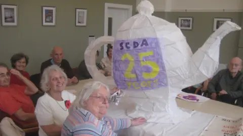 Bedminster Winter Lanterns  A group of older people in a large community room sitting round a table with a large teapot-shaped white lantern on it