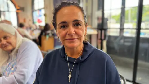 Morag Burnard has dark hair tied back. She is wearing a navy sweatshirt and silver pendant necklace - and sitting near the doors at a busy cafe
