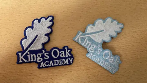 Cabot Learning Federation Two embroidered iron-on school badges lie on a light wooden surface. Both feature the words “King’s Oak Academy” beneath a large oak leaf, with one badge outlined in dark blue and the other in a pale blue colour.