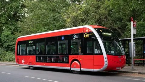 File image of one of the new 358 red single-decker buses, which have a slanted windscreen that curves under at the bonnet, lower windows for some seats, and red hubcaps over the tyres. The pantograph arm is not visible