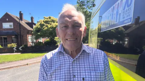 A man in his late seventies wearing a white and blue checked shirt, standing next to an ambulance.