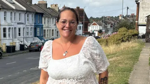 BBC Kelly Warwick standing outside her house in Brighton. She wears a white blouse and dark jeans. She has dark hair tied up, and glasses.