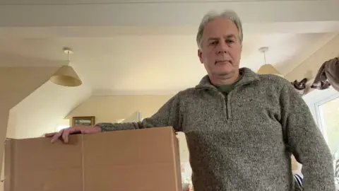 Marcus Kats. He is wearing a grey jumper and has his arm outstretched on a cardboard box.