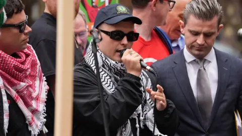 PA Three men stand in a line. One holds a mic up. The two men on the left wear scarves and the one on the right wears a grey suit and silver tie. 