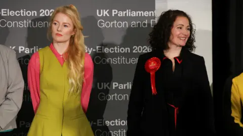 Reuters Green Party candidate Hannah Spencer looks serious as she stands next to Labour Party candidate Angeliki Stogia, who smiles, on stage before results are announced.