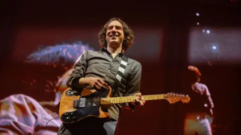 Debbie Hickey/Getty Images Snow Patrol Performs At 3Arena, Dublin.  Lead singer Gary Lightbody is holding an electric guitar and smiling.  He has wavy, greying dark hair and is wearing a grey shirt and jeans. He also appears on large TV screens at the back of the stage. 