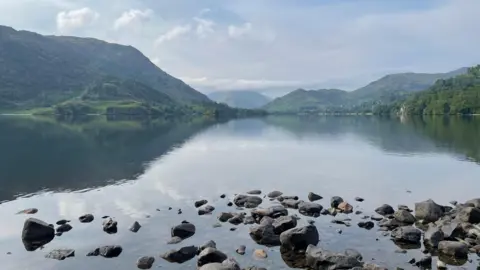 Georgia Roper A large lake which has a collection of rocks in the foreground. There are hills in the distance which are covered in grass. The water is calm with reflections of the hills visible. The sky is covered in white clouds with bits of blue peeking through.
