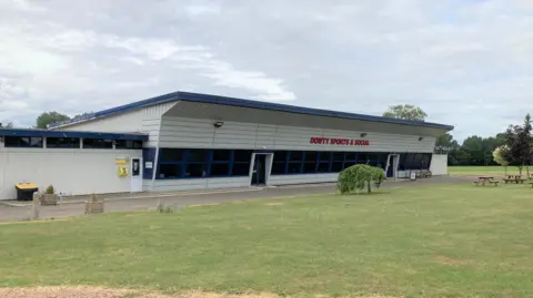 A large grey building with a slanting roof. The words "Dowty Sports & Social" are written in large red letters above the entrance. Grass surrounds the building and there are several picnic benches and trees in the distance.