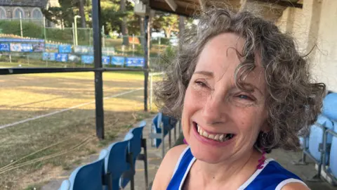 Debby Burnside - a woman with grey short curly hair who is wearing a blue running top, sitting on a chair beside a rugby pitch smiling.